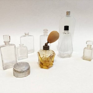 May include: A collection of vintage glass perfume bottles with various shapes and sizes. The bottles are clear glass and some have stoppers. There is also a small, round, silver-colored container.