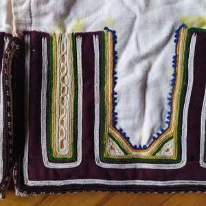 May include: Close-up of a white fabric garment with a burgundy trim and intricate embroidery in shades of white, green, yellow, and blue.