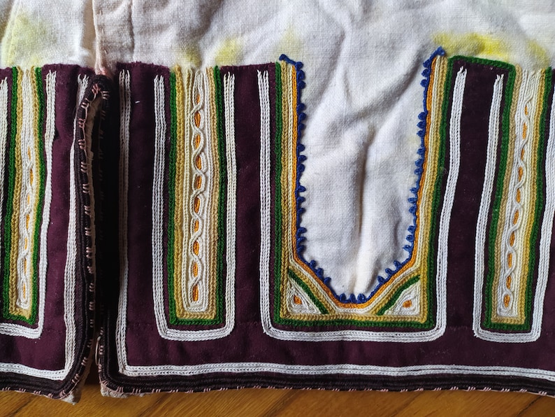 May include: Close-up of a white fabric garment with a burgundy trim and intricate embroidery in shades of white, green, yellow, and blue.