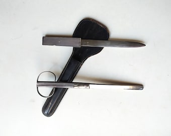 Op de afbeelding: Een set zilverkleurige briefopener en schaar met een zwarte leren schede. De briefopener is een enkele mes met een zilveren handvat. De schaar heeft een rond handvat en een lange mes.