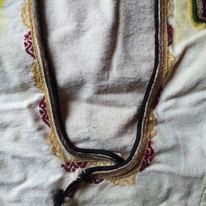 May include: Close-up of a white fabric with a decorative embroidered neckline. The neckline features a black, brown, and gold trim with a red and gold pattern.