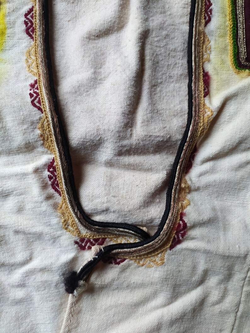 May include: Close-up of a white fabric with a decorative embroidered neckline. The neckline features a black, brown, and gold trim with a red and gold pattern.