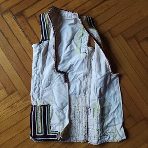 May include: A white waistcoat with black, green and purple trim. The waistcoat is made from a lightweight fabric and has a simple design.