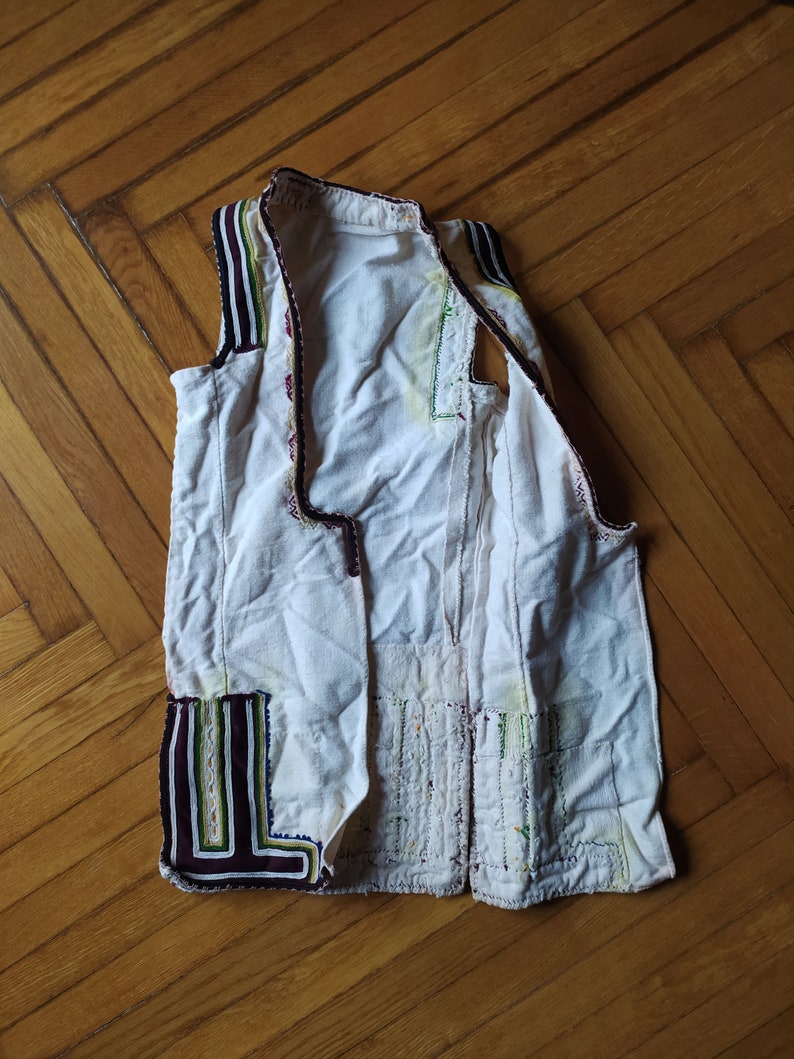 May include: A white waistcoat with black, green and purple trim. The waistcoat is made from a lightweight fabric and has a simple design.
