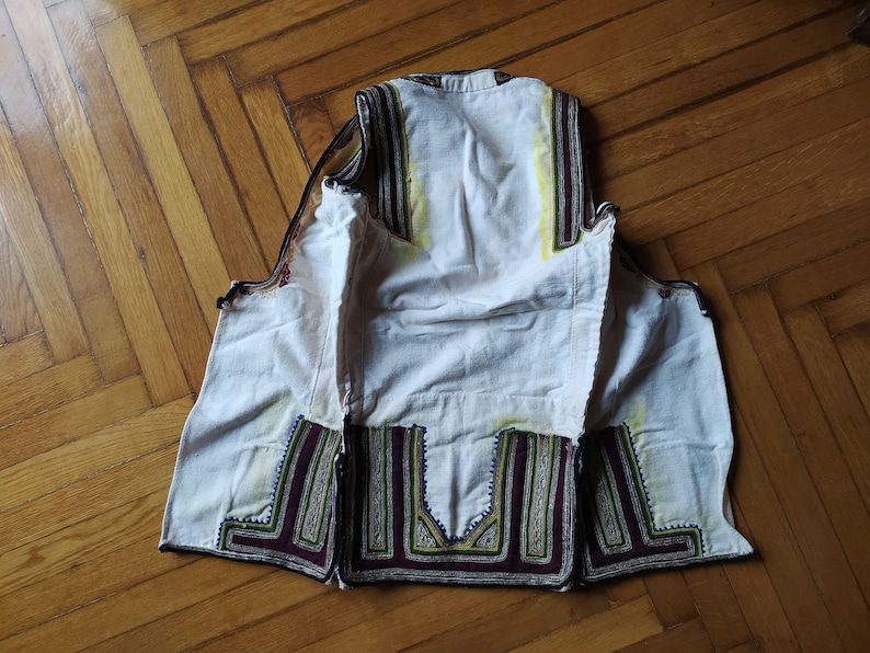 May include: A white waistcoat with intricate embroidery in shades of green, brown, and purple. The waistcoat is lined with white fabric and has a simple, classic design.