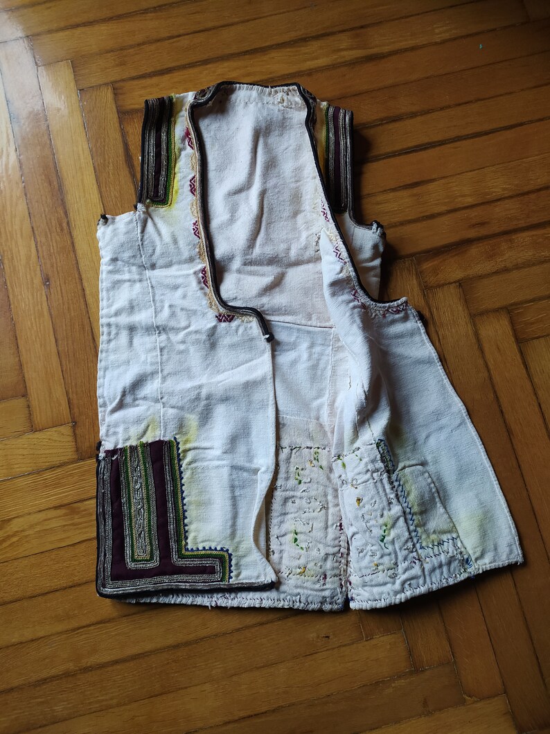 May include: A white sleeveless waistcoat with intricate embroidery in shades of red, green, yellow, and black. The waistcoat has a button closure and is made of a lightweight fabric.