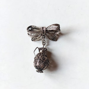 Op de afbeelding: Zilverkleurige filigraan broche met een strikdesign aan de bovenkant en een kleine kruikhanger eronder. De broche heeft ingewikkelde open patronen en staat tegen een witte achtergrond.
