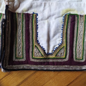 May include: Close-up of a white fabric garment with intricate embroidery in shades of green, gold, and silver. The embroidery is in a geometric pattern and is accented with a dark burgundy fabric.
