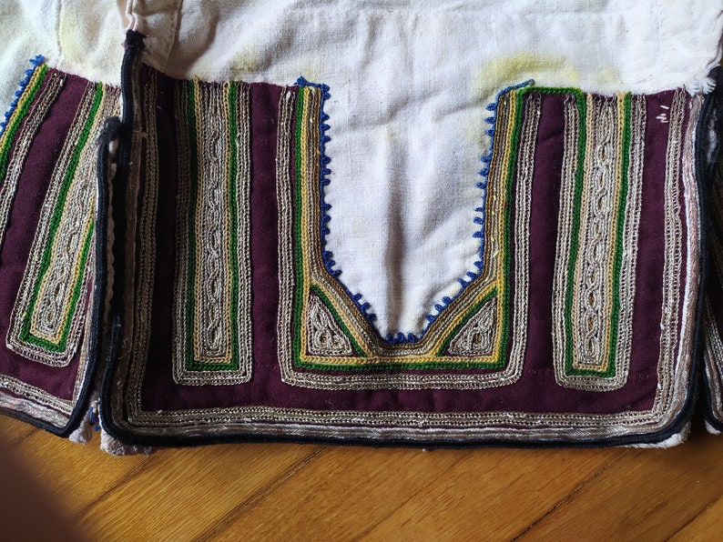 May include: Close-up of a white fabric garment with intricate embroidery in shades of green, gold, and silver. The embroidery is in a geometric pattern and is accented with a dark burgundy fabric.
