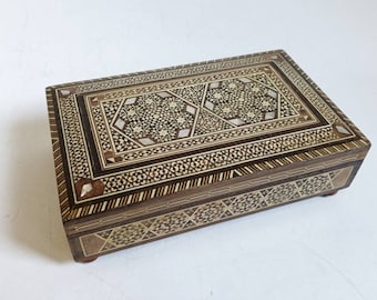 May include: A rectangular wooden box with intricate inlaid patterns in shades of brown and white. The box has a hinged lid and small feet on the bottom.