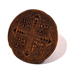 May include: A round, wooden object with intricate geometric carvings. The object is brown and features a central cross design surrounded by repeating patterns and text. The overall aesthetic suggests a historical or cultural artifact.