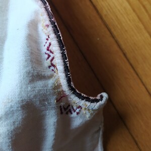 May include: Close-up of a white fabric with a black, red, and gold embroidered trim. The fabric is folded over and the trim is visible on both sides.