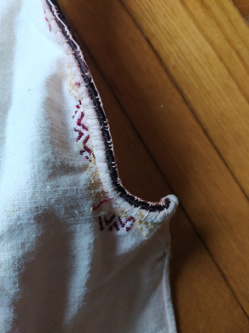 May include: Close-up of a white fabric with a black, red, and gold embroidered trim. The fabric is folded over and the trim is visible on both sides.