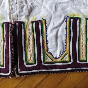 May include: Close-up of a white fabric garment with intricate embroidery. The embroidery features a combination of white, green, yellow, and blue threads, creating a geometric pattern. The fabric is edged with a dark purple trim.