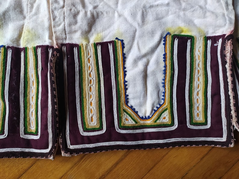 May include: Close-up of a white fabric garment with intricate embroidery. The embroidery features a combination of white, green, yellow, and blue threads, creating a geometric pattern. The fabric is edged with a dark purple trim.