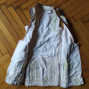 May include: A white sleeveless vest with a decorative pattern of colourful stitching on the front. The vest has a brown and black striped trim around the neck and armholes.