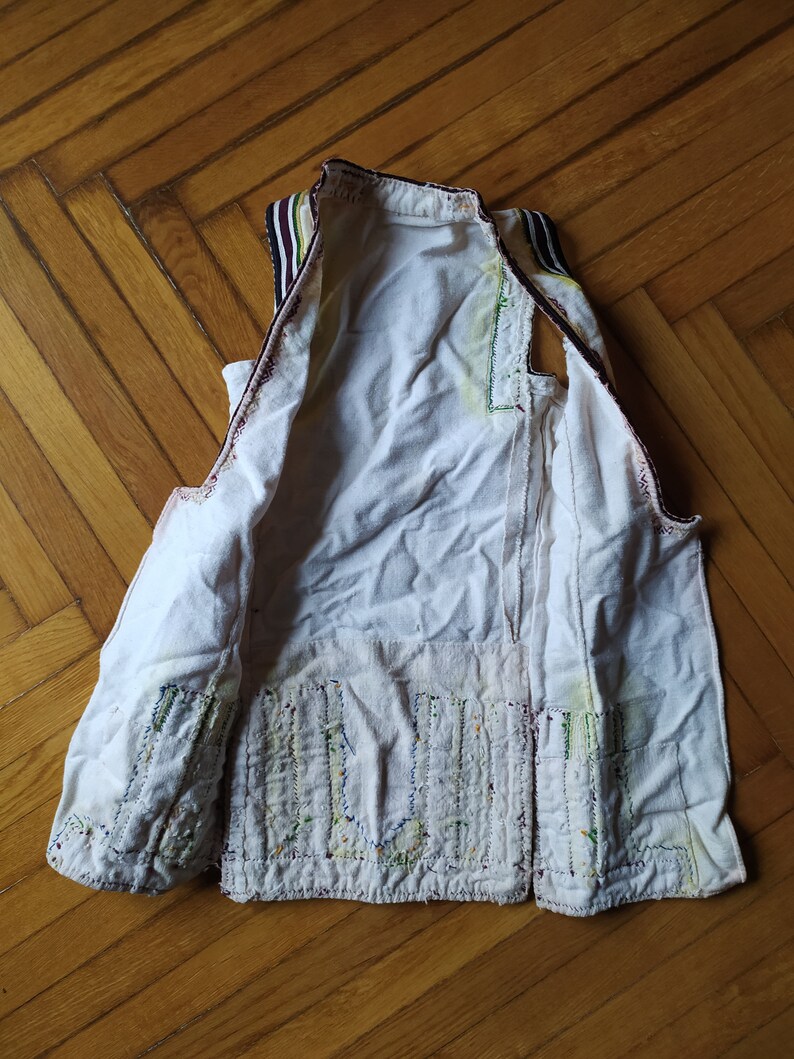 May include: A white sleeveless vest with a decorative pattern of colourful stitching on the front. The vest has a brown and black striped trim around the neck and armholes.