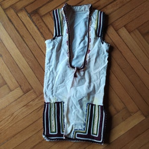 May include: A white sleeveless waistcoat with colourful embroidery around the neckline and hem. The waistcoat has a traditional folk design.