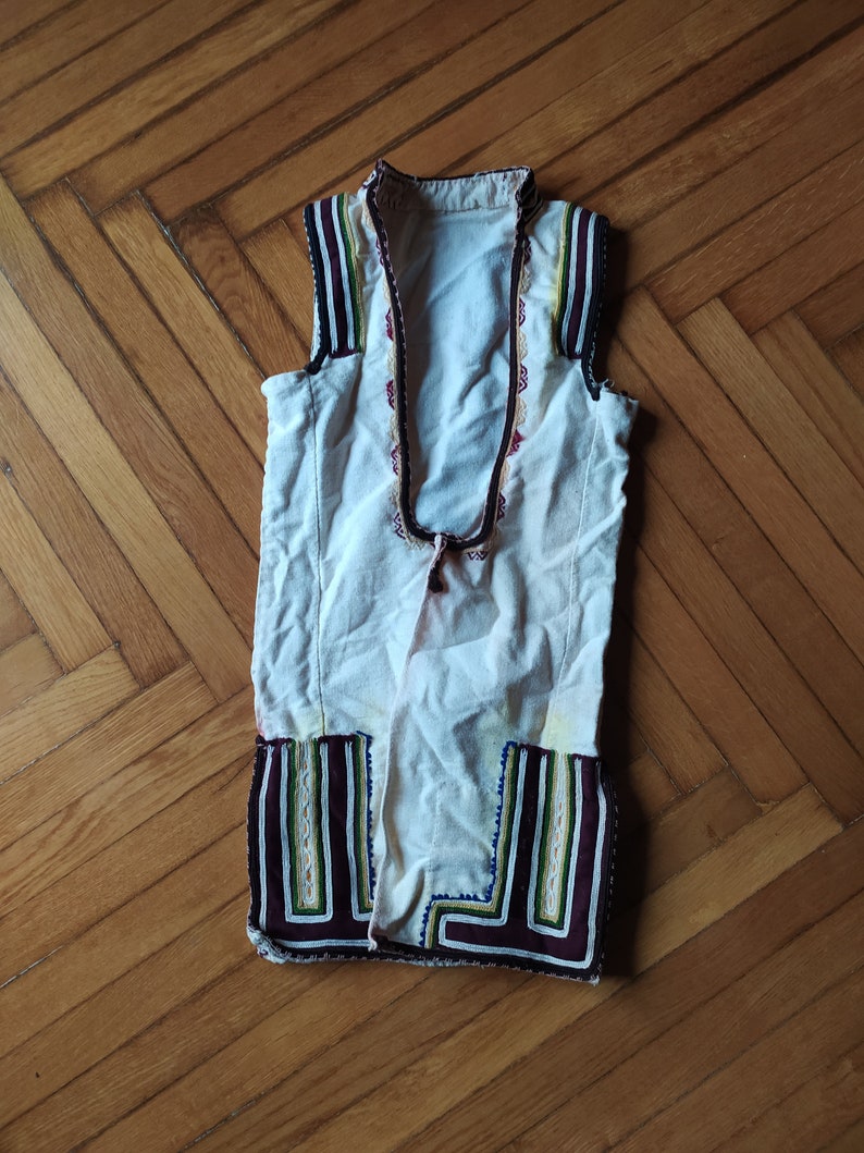 May include: A white sleeveless waistcoat with colourful embroidery around the neckline and hem. The waistcoat has a traditional folk design.