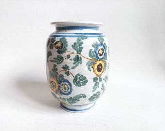 May include: A white ceramic vase with a floral design. The vase has a round body and a narrow neck. The flowers are painted in shades of blue, yellow, and green.