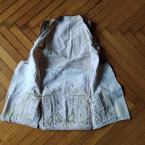 May include: A white waistcoat with colourful embroidery on the front and back. The waistcoat is turned inside out, showing the lining and stitching.