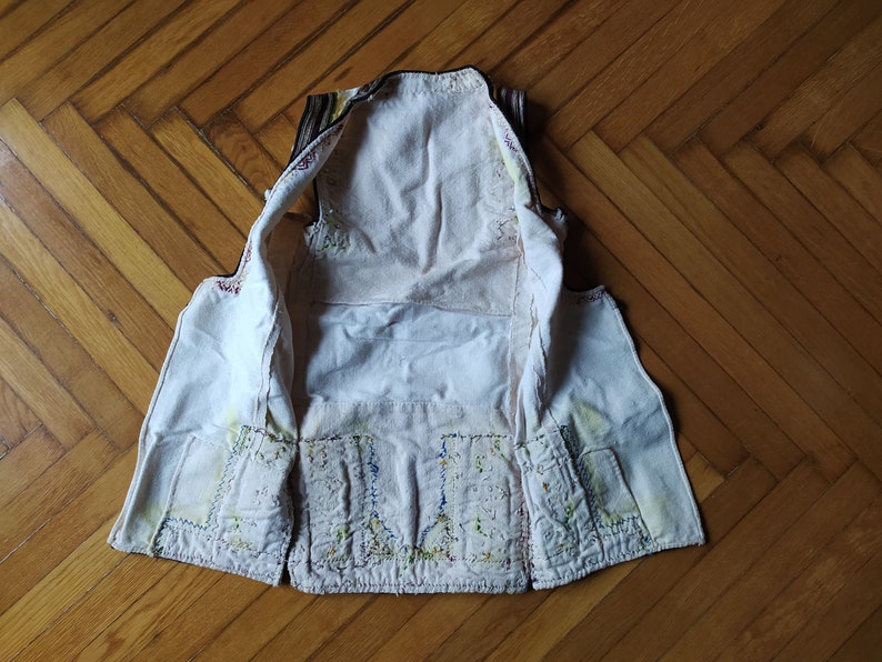 May include: A white waistcoat with colourful embroidery on the front and back. The waistcoat is turned inside out, showing the lining and stitching.