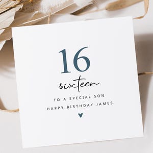 May include: A white square birthday card with the number "16" in navy blue. The word "sixteen" is written below in cursive. The text "TO A SPECIAL SON HAPPY BIRTHDAY JAMES" is below, with a small heart.
