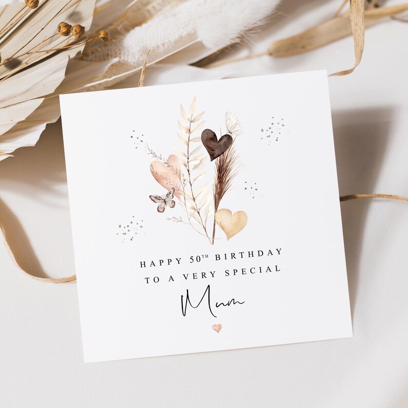 Pode incluir: Um cart&atilde;o de felicita&ccedil;&otilde;es branco quadrado com o texto "HAPPY 50TH BIRTHDAY TO A VERY SPECIAL Mum". Apresenta ilustra&ccedil;&otilde;es em aquarela de cora&ccedil;&otilde;es, uma borboleta e flores secas em tons de castanho, rosa e bege.