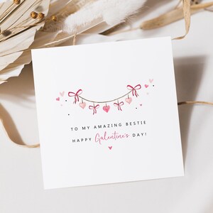 Puede incluir: Una tarjeta blanca con una guirnalda de corazones y lazos rosas. El texto dice "TO MY AMAZING BESTIE" y "HAPPY Galentine's DAY!" en escritura rosa. Peque&ntilde;os puntos y corazones negros est&aacute;n esparcidos alrededor de la guirnalda.