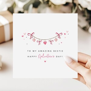 Puede incluir: Una tarjeta de felicitaci&oacute;n blanca con el texto "TO MY AMAZING BESTIE HAPPY Galentine's DAY!" en escritura negra y rosa. La tarjeta presenta una guirnalda de corazones y lazos rosas. Una mano sostiene la tarjeta.