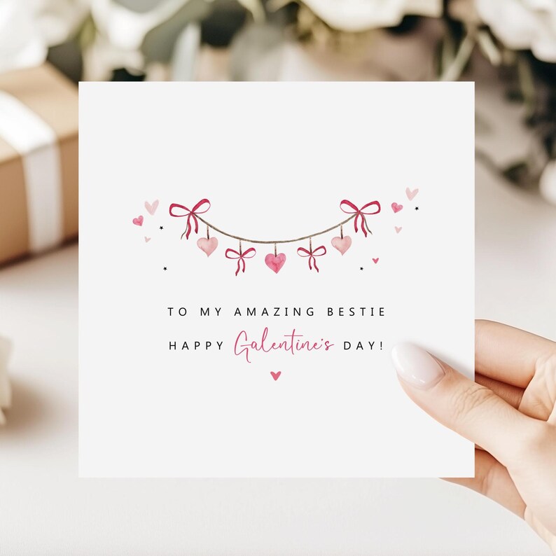 Puede incluir: Una tarjeta de felicitaci&oacute;n blanca con el texto "TO MY AMAZING BESTIE HAPPY Galentine's DAY!" en escritura negra y rosa. La tarjeta presenta una guirnalda de corazones y lazos rosas. Una mano sostiene la tarjeta.