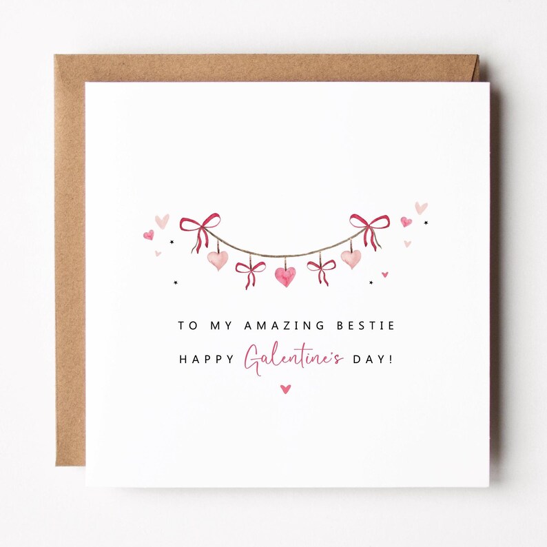 Puede incluir: Tarjeta de felicitaci&oacute;n cuadrada con fondo blanco y sobre marr&oacute;n. La tarjeta presenta una guirnalda de corazones y lazos rosas. El texto dice "TO MY AMAZING BESTIE" y "HAPPY Galentine's DAY!"