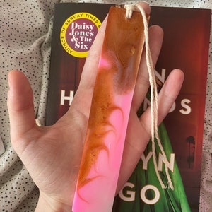 May include: Pink and gold resin bookmark with a white tassel. The bookmark is rectangular and has a marbled design.