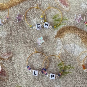 May include: A set of eight gold hoop earrings with white letter beads and pearl accents. The letters spell out the words "H.S", "N.H", "Z.M", "L.T", "L.P", and "I.D".