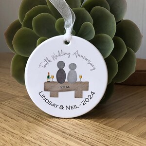 May include: White ceramic ornament with a gray illustration of two people sitting on a bench with a bottle of champagne and glasses. The text "Tenth Wedding Anniversary 2014 - 2024 Lindsay & Neil" is printed on the ornament.