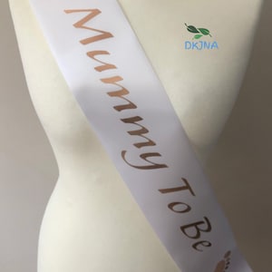 May include: White satin sash with gold foil lettering that reads "Mommy To Be" and gold foil baby footprints on each end.