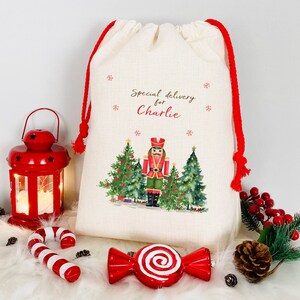 May include: A white canvas gift bag with red drawstring, featuring a watercolour illustration of a nutcracker standing in front of Christmas trees. The bag is personalised with the text "Special delivery for Charlie".