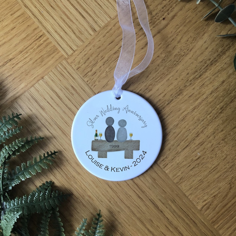 May include: White ceramic ornament with a silver wedding anniversary design. It features two figures sitting on a bench with a bottle of champagne and glasses. The text reads "Silver Wedding Anniversary Louise & Kevin - 2024" and "1999".