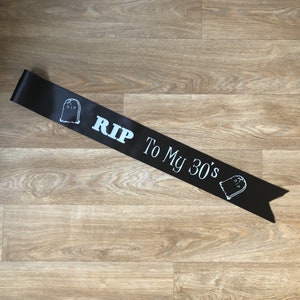 May include: Black satin sash with white text and tombstone graphics. The text reads "RIP To My 30's". A fun accessory for a milestone birthday celebration. The sash is laid on a light brown wooden surface.