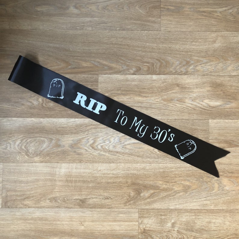 May include: Black satin sash with white text and tombstone graphics. The text reads "RIP To My 30's". A fun accessory for a milestone birthday celebration. The sash is laid on a light brown wooden surface.