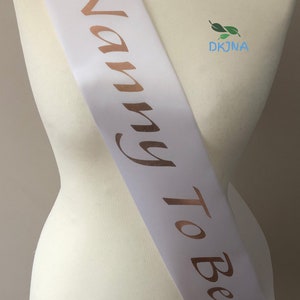 May include: White satin sash with the words "Nanny To Be" in gold lettering. The sash has a small bird design at each end.