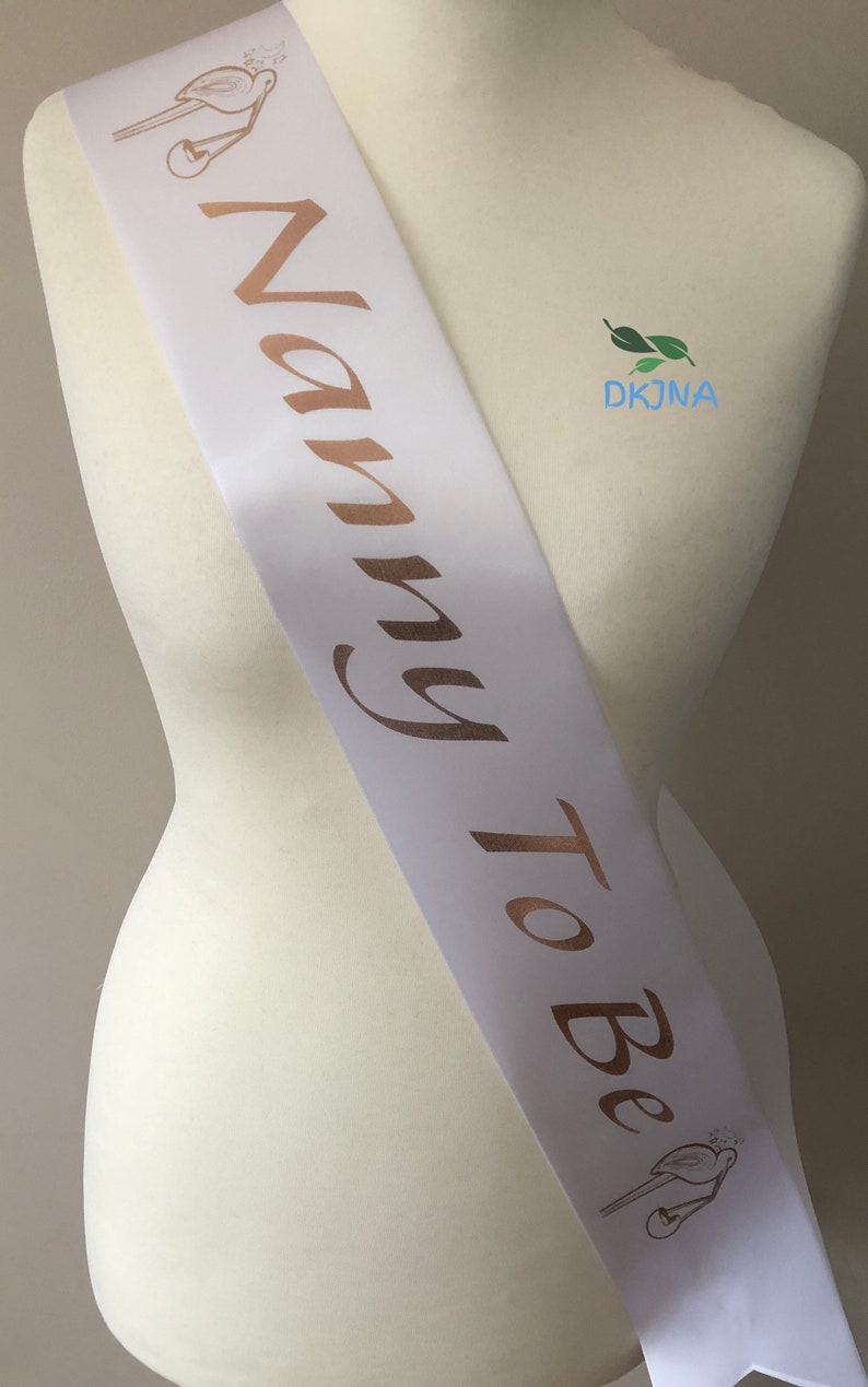 May include: White satin sash with the words "Nanny To Be" in gold lettering. The sash has a small bird design at each end.