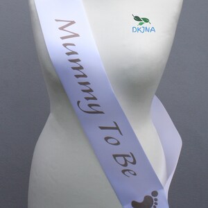 May include: White satin sash with the text "Mummy To Be" in brown script. The sash has two small footprints on either end.