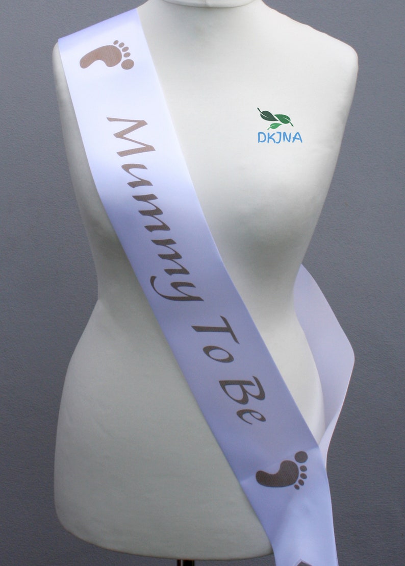 May include: White satin sash with the text "Mummy To Be" in brown script. The sash has two small footprints on either end.