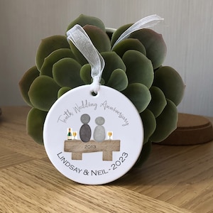 May include: White ceramic ornament with a gray illustration of a couple sitting on a bench with glasses of champagne. The text "Tenth Wedding Anniversary 2013 Lindsay & Neil - 2023" is printed on the ornament.