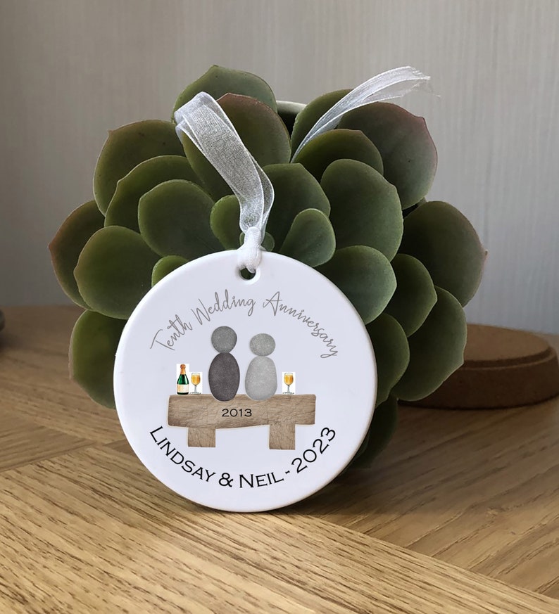 May include: White ceramic ornament with a gray illustration of a couple sitting on a bench with glasses of champagne. The text "Tenth Wedding Anniversary 2013 Lindsay & Neil - 2023" is printed on the ornament.