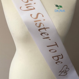 May include: White satin sash with gold foil lettering that reads "Big Sister To Be" and two gold foil hearts on each end.