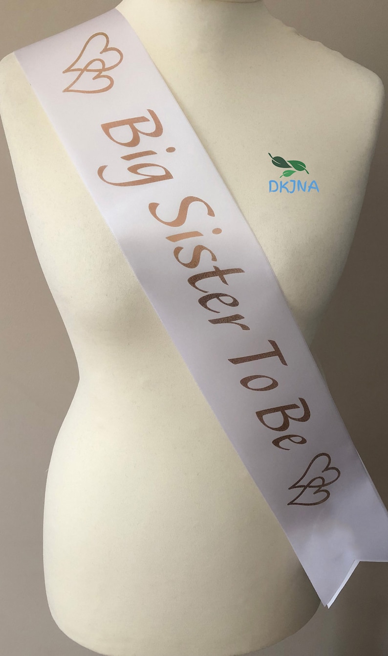 May include: White satin sash with gold foil lettering that reads "Big Sister To Be" and two gold foil hearts on each end.