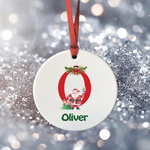 May include: White ceramic Christmas bauble featuring a red letter O, a Santa Claus illustration, and the name Oliver in green text.