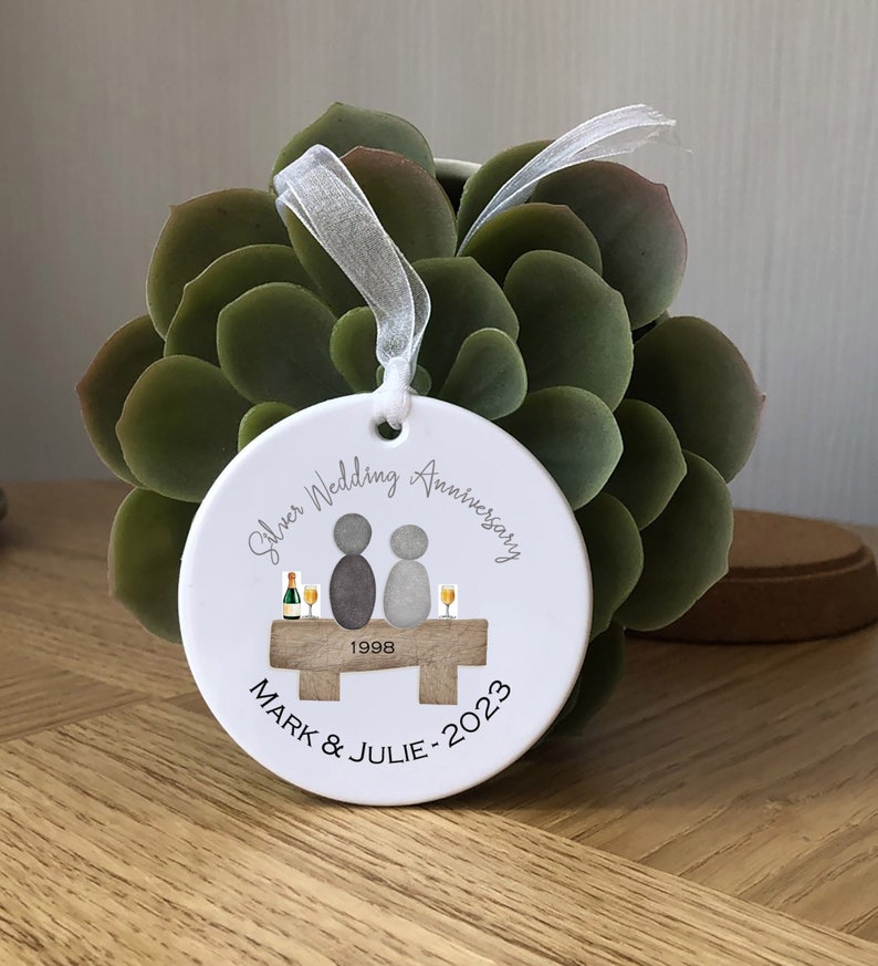 May include: White round ceramic ornament with a silver wedding anniversary design. It features two figures sitting on a bench with a bottle of champagne and glasses. The text reads "Silver Wedding Anniversary 1998 MARK & JULIE - 2023".